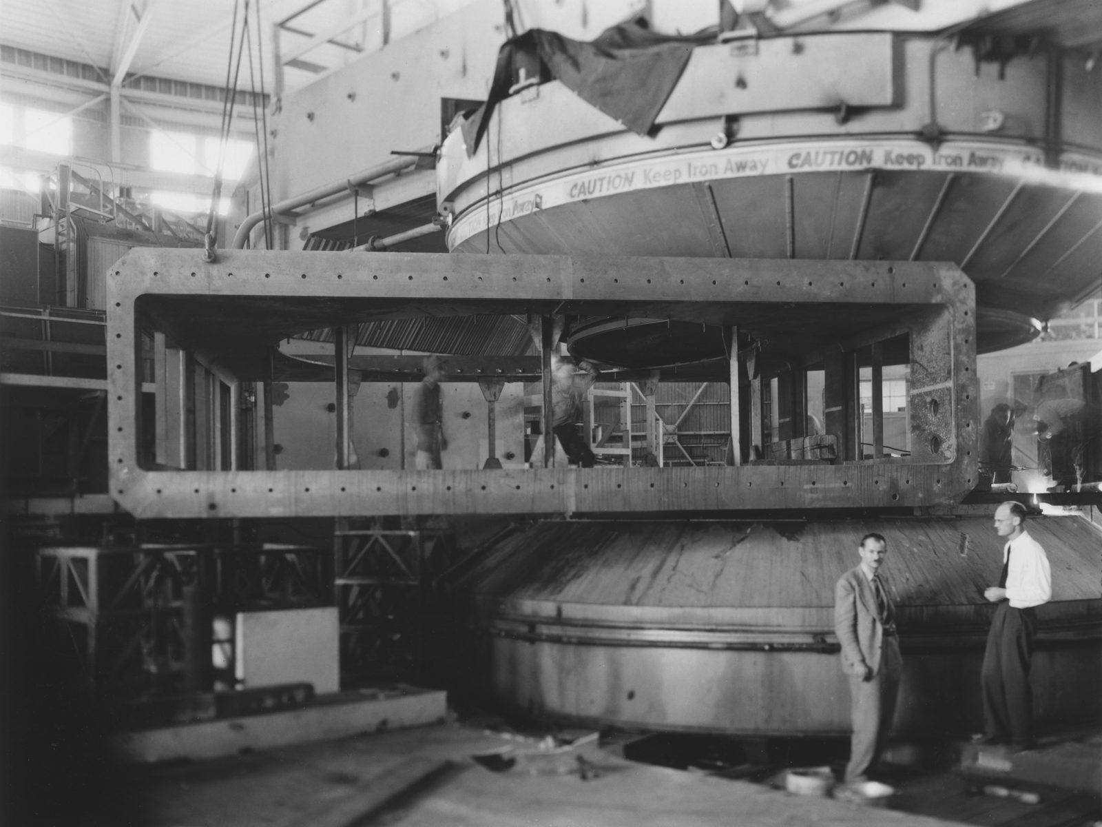 A rectangular steel vacuum tank the size of a small room hangs from crane cables between the enormous upper and lower poles of the 184-inch magnet, its upper coil tank lettered CAUTION KEEP IRON AWAY; two men stand beside the lower pole.