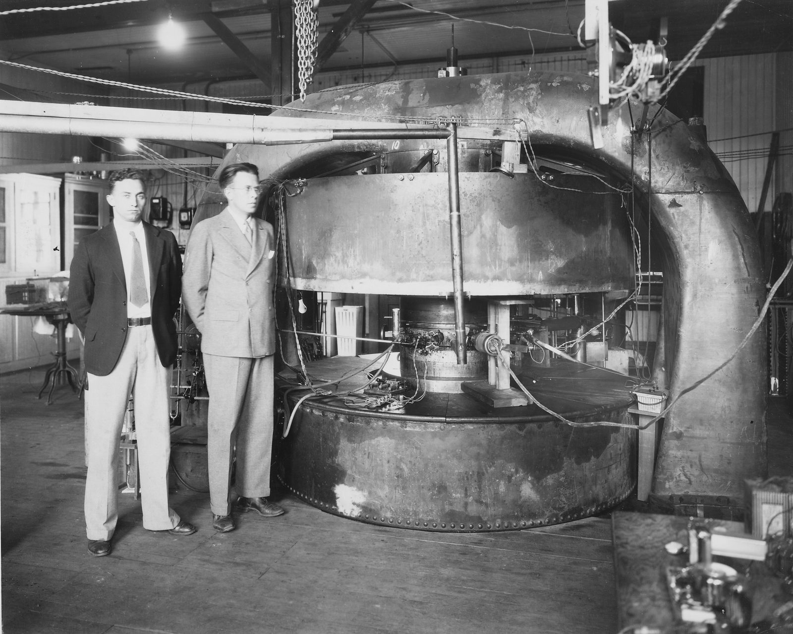 Two men in suits stand beside a large electromagnet whose round pole pieces, taller than they are, close on a flat vacuum chamber crossed with wires and glass tubing, in a wooden laboratory building.