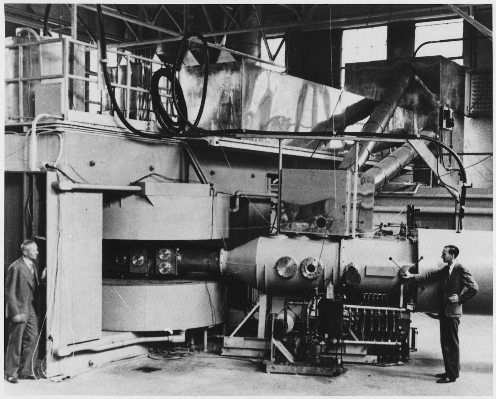 A large cyclotron in a high-ceilinged hall: a boxy magnet yoke at left with coil tanks above and below the chamber, a long tapered beam pipe running right to a cylindrical tank with ports, and a man standing at each end for scale.