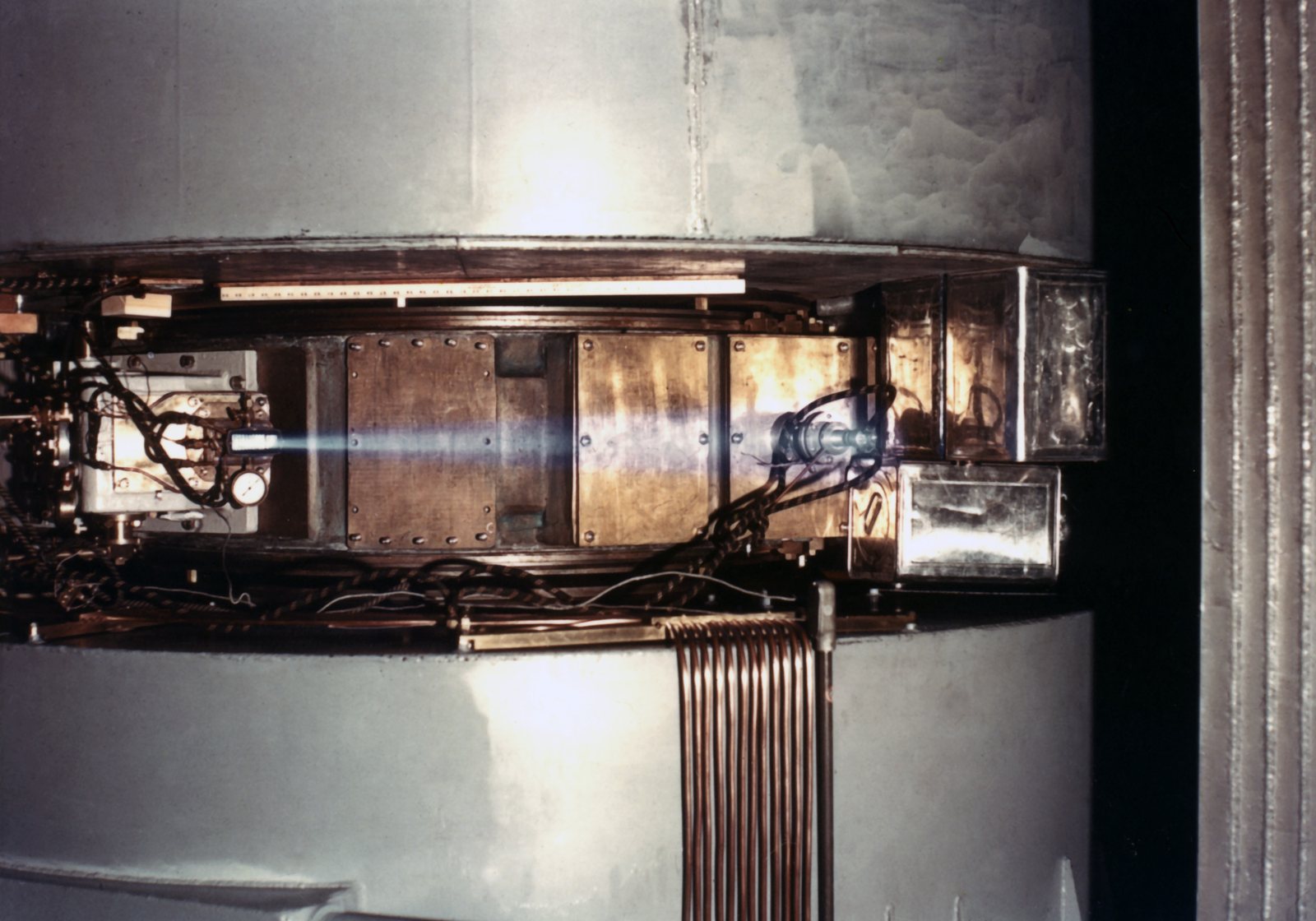 Color photograph: a brilliant blue-white beam emerges horizontally from a brass port on the side of the 60-inch cyclotron's chamber and crosses open air between the magnet's grey pole housings; copper cooling tubes hang below.