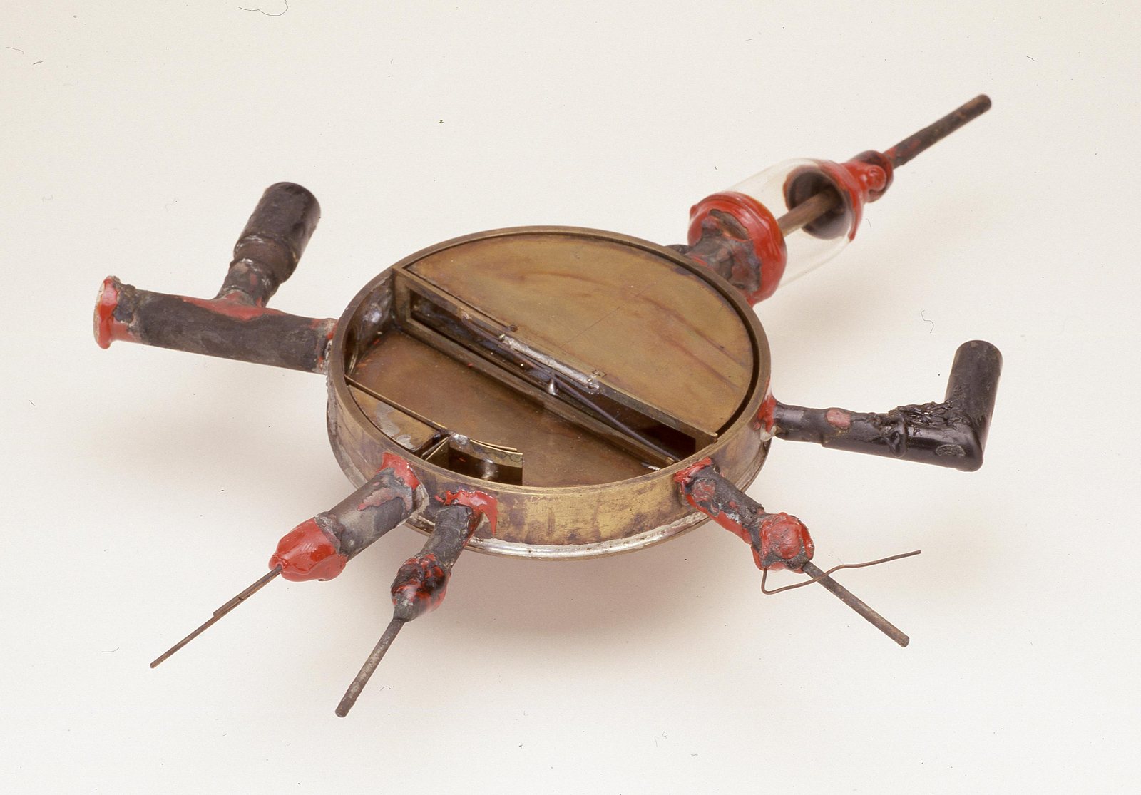 The original first cyclotron chamber: a shallow round brass can a few inches across with its lid off, one hollow D-shaped brass electrode filling half the interior, and six stubby tubes radiating from the rim, each sealed with red wax; one carries a small glass insulator.