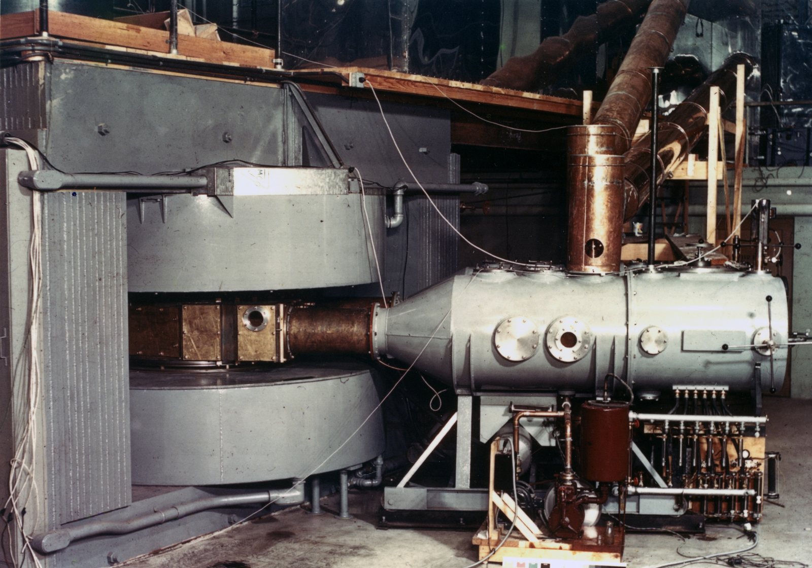 A 1939 cyclotron in a laboratory hall: a grey magnet with round coil tanks above and below the chamber at left, a tapered section leading right to a long cylindrical vacuum tank with round ports, pumps and copper piping beneath and beside it.